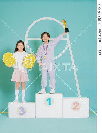 Two children stand on a podium after winning a competition. One child holds a trophy, while the other displays pom poms. The setting is bright and colorful, highlighting their achievements. Two children stand on a podium after winning a competition. One child holds a trophy, while the other displays pom poms. The setting is bright and colorful, highlighting their achievements. 139259729