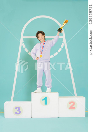 A child stands proudly on the first place podium holding a trophy. The child wears a medal and a pink outfit. The setting shows a simple and fun background with bright colors. A child stands proudly on the first place podium holding a trophy. The child wears a medal and a pink outfit. The setting shows a simple and fun background with bright colors. 139259731