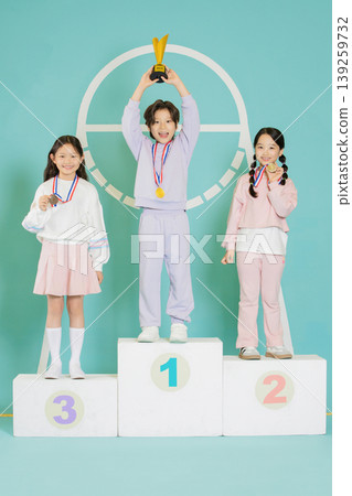 Three children stand on a podium after a sports event. One child holds a trophy on the first place spot, while others show their medals. They smile in a gym setting. 139259732