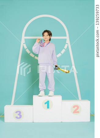 A young athlete stands proudly on a podium after winning a competition. They hold a trophy in one hand and touch their medal with the other. The background has a large hoop design. A young athlete stands proudly on a podium after winning a competition. They hold a trophy in one hand and touch their medal with the other. The background has a large hoop design. 139259733