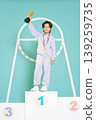 A young child stands proudly on the first place podium holding a trophy high. The child wears a medal around the neck and is dressed in a simple outfit. 139259735