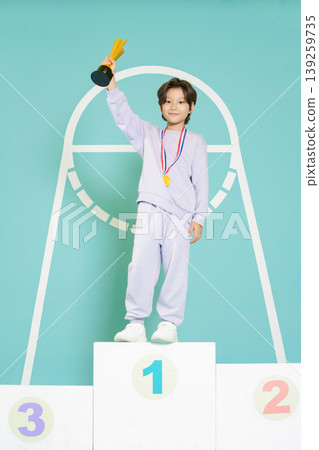 A young child stands proudly on the first place podium holding a trophy high. The child wears a medal around the neck and is dressed in a simple outfit. A young child stands proudly on the first place podium holding a trophy high. The child wears a medal around the neck and is dressed in a simple outfit. 139259735