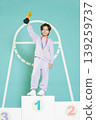 A child stands proudly on the first place podium holding a trophy. The child wears a medal and sportswear. The background is simple and bright, highlighting the achievement. 139259737