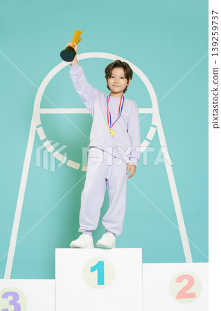 A child stands proudly on the first place podium holding a trophy. The child wears a medal and sportswear. The background is simple and bright, highlighting the achievement. A child stands proudly on the first place podium holding a trophy. The child wears a medal and sportswear. The background is simple and bright, highlighting the achievement. 139259737