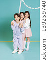 Three kids stand together, smiling for the camera. They wear casual clothes in bright colors. The background is simple and colorful, creating a lively scene. The kids show joy in their friendship. 139259740