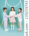 Three kids stand on a studio floor with a light blue backdrop. They wear colorful casual outfits. Each child places their hands together for a playful moment. 139259743