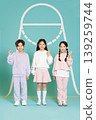 Three children are standing together in a bright room with a blue background. They wear colorful and comfortable clothes. 139259744