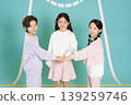Three children stand in a playful setting, smiling at the camera while joining hands. They are dressed in colorful clothes and look happy together. The background adds a fun touch. 139259746