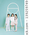 Three children stand side by side in a studio. They wear matching outfits in light colors. The background has a simple design. The setting is bright and welcoming. 139259749