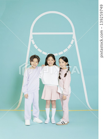 Three children stand side by side in a studio. They wear matching outfits in light colors. The background has a simple design. The setting is bright and welcoming. 139259749