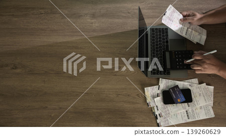 Hands using calculator on a laptop with receipts and credit card on a wooden desk, managing personal expenses and budgeting in a home setting and tax preparation content. 139260629