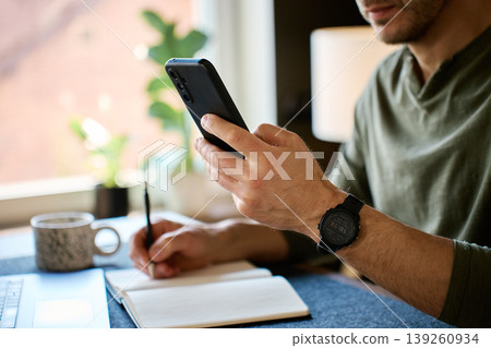 Man checking smartphone while writing notes at home desk Man checking smartphone while writing notes at home desk 139260934