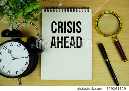 Crisis Ahead text written on notepad with alarm clock and magnifying glass flat lay on wooden background Crisis Ahead text written on notepad with alarm clock and magnifying glass flat lay on wooden background 139261129