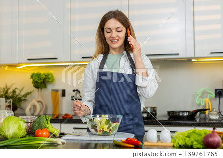 Middle-aged woman smiling during smartphone call while mixing fresh vegetable salad in home kitchen Middle-aged woman smiling during smartphone call while mixing fresh vegetable salad in home kitchen 139261765