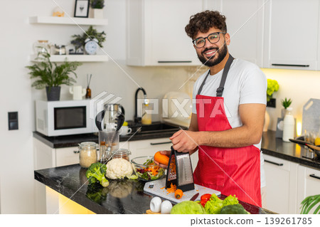 Arabian young man grating carrot in kitchen at home for fresh salad meal prep and healthy nutrition Arabian young man grating carrot in kitchen at home for fresh salad meal prep and healthy nutrition 139261785