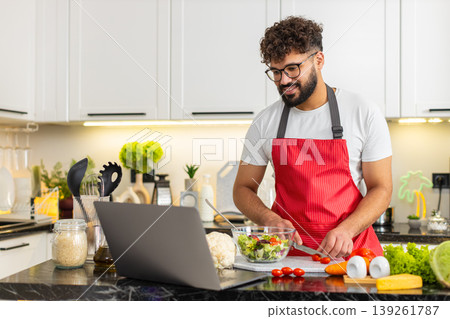 Arabian young adult man cooking fresh salad in kitchen watching recipe on laptop for quick meal prep Arabian young adult man cooking fresh salad in kitchen watching recipe on laptop for quick meal prep 139261787