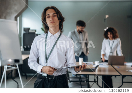 Man is standing and holding notepad. Group of people are working in the office together as a team Man is standing and holding notepad. Group of people are working in the office together as a team 139262119