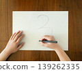 A child's hand writing the number 3 with a black pen on white paper; the child is learning at a wooden table. 139262335