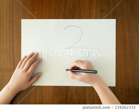 A child's hand writing the number 3 with a black pen on white paper; the child is learning at a wooden table. 139262335