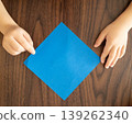 A top-down view of a child's hands placing blue origami paper on a wooden table. 139262340