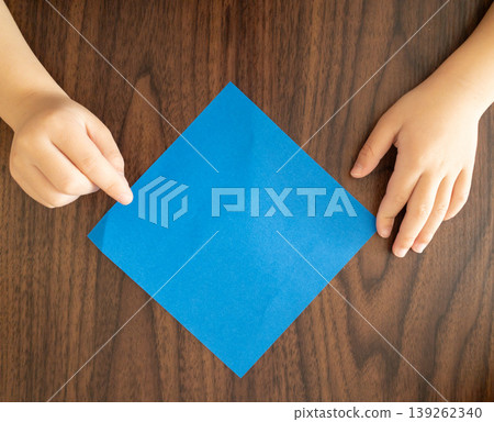 A top-down view of a child's hands placing blue origami paper on a wooden table. 139262340