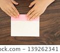A child's hand folding pink origami paper on a wooden table. 139262341