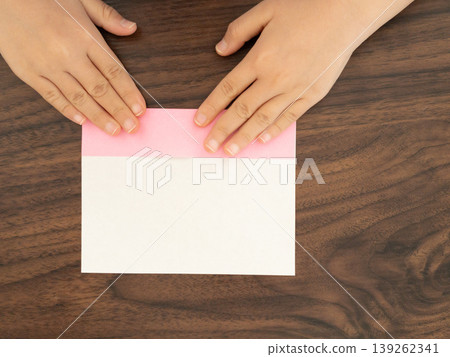 A child's hand folding pink origami paper on a wooden table. 139262341