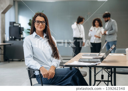Woman is sitting and looking at the camera. Group of people are working in the office together as a team Woman is sitting and looking at the camera. Group of people are working in the office together as a team 139262348