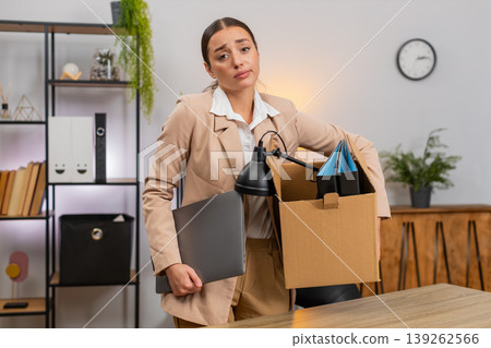 Young businesswoman packs items into cardboard box takes laptop looks back at office with farewell Young businesswoman packs items into cardboard box takes laptop looks back at office with farewell 139262566