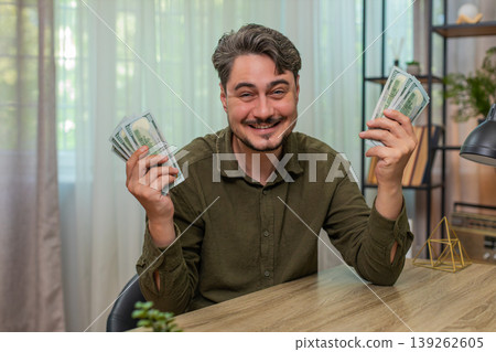 Middle-aged man counts cash smiling showing big financial success dollar bills wealth at table desk Middle-aged man counts cash smiling showing big financial success dollar bills wealth at table desk 139262605