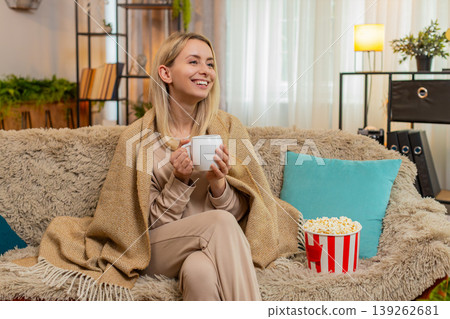 Adult woman wrapped in blanket sitting on sofa with cup of tea watching series thoughtfully at home Adult woman wrapped in blanket sitting on sofa with cup of tea watching series thoughtfully at home 139262681
