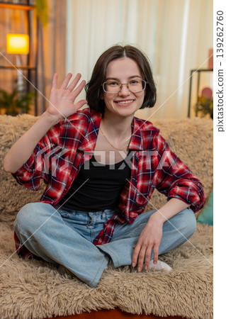 Young woman smiling and waving hand at home, greeting someone with friendly and warm expression 139262760