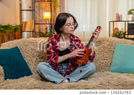 Young woman playing ukulele at home, focused on learning chord and smiling with satisfaction and joy Young woman playing ukulele at home, focused on learning chord and smiling with satisfaction and joy 139262763