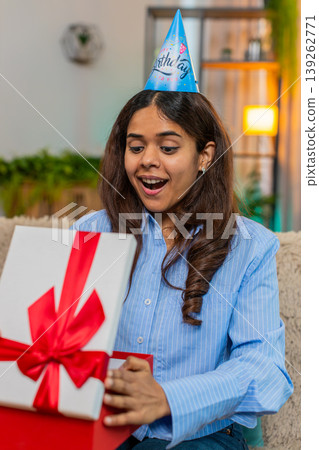 Indian woman opening birthday gift on home sofa reacting with surprise and joy smiling with delight Indian woman opening birthday gift on home sofa reacting with surprise and joy smiling with delight 139262771