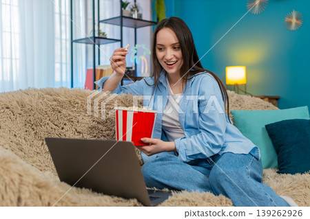 Woman happily eating popcorn watching movie on laptop at home sofa emotionally laughing enjoyment Woman happily eating popcorn watching movie on laptop at home sofa emotionally laughing enjoyment 139262926