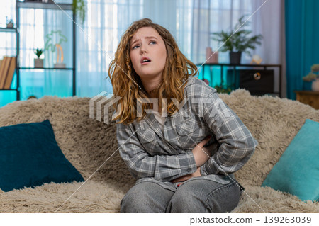 Woman sitting on sofa holding stomach suffering from pain and discomfort feeling unwell at home Woman sitting on sofa holding stomach suffering from pain and discomfort feeling unwell at home 139263039