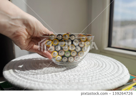 A young woman places a glass bowl filled with used batteries onto a wooden shelf for future disposal. This process demonstrates conscious household waste sorting and care for the environment. A young woman places a glass bowl filled with used batteries onto a wooden shelf for future disposal. This process demonstrates conscious household waste sorting and care for the environment. 139264726