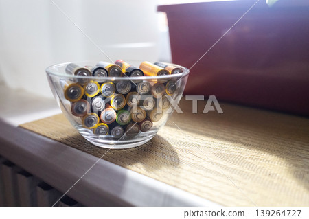 A young woman places a glass bowl filled with used batteries onto a wooden shelf for future disposal. This process demonstrates conscious household waste sorting and care for the environment. A young woman places a glass bowl filled with used batteries onto a wooden shelf for future disposal. This process demonstrates conscious household waste sorting and care for the environment. 139264727