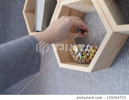 A young woman places a glass bowl filled with used batteries onto a wooden shelf for future disposal. This process demonstrates conscious household waste sorting and care for the environment. A young woman places a glass bowl filled with used batteries onto a wooden shelf for future disposal. This process demonstrates conscious household waste sorting and care for the environment. 139264733