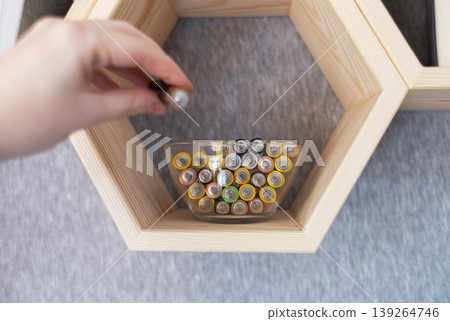 A young woman places a glass bowl filled with used batteries onto a wooden shelf for future disposal. This process demonstrates conscious household waste sorting and care for the environment. 139264746