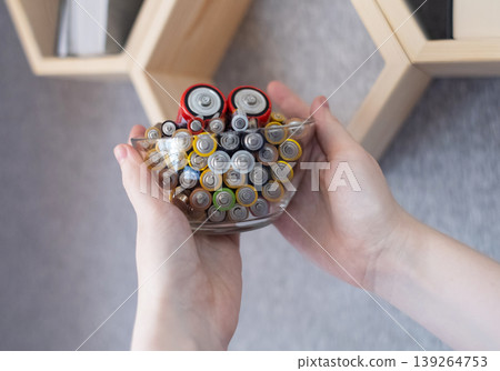 A young woman places a glass bowl filled with used batteries onto a wooden shelf for future disposal. This process demonstrates conscious household waste sorting and care for the environment. A young woman places a glass bowl filled with used batteries onto a wooden shelf for future disposal. This process demonstrates conscious household waste sorting and care for the environment. 139264753