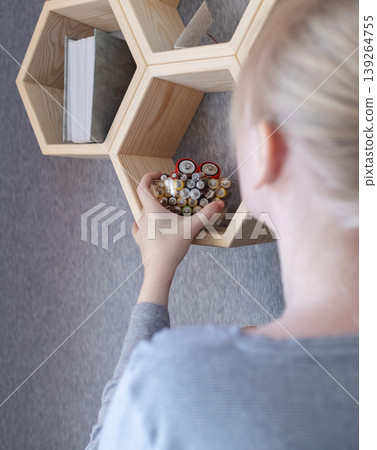 A young woman places a glass bowl filled with used batteries onto a wooden shelf for future disposal. This process demonstrates conscious household waste sorting and care for the environment. A young woman places a glass bowl filled with used batteries onto a wooden shelf for future disposal. This process demonstrates conscious household waste sorting and care for the environment. 139264755