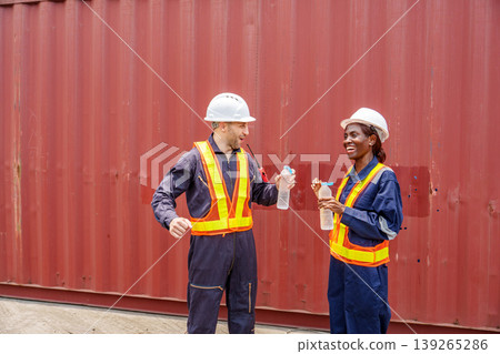 Smiling mid-adult Caucasian male and Black female industrial workers sharing a laugh while drinking water at an Asian container yard. Smiling mid-adult Caucasian male and Black female industrial workers sharing a laugh while drinking water at an Asian container yard. 139265286
