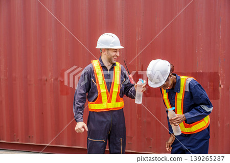 Smiling mid-adult Caucasian male and Black female industrial workers sharing a laugh while drinking water at an Asian container yard. 139265287