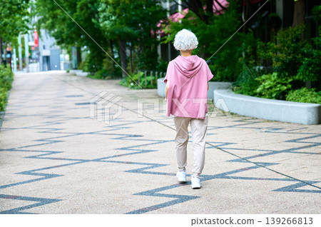 一位老婦人背對著鏡頭，走在綠樹成蔭的街道上。 139266813