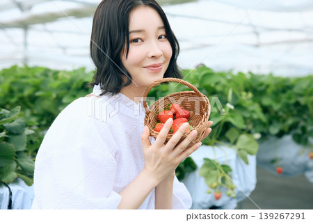 A young woman with a smile enjoying strawberry picking; a Japanese woman holding a basket of freshly picked strawberries in a greenhouse. 139267291