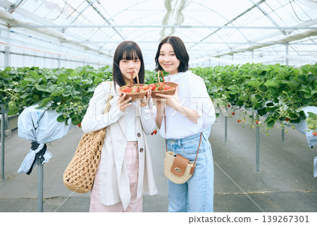 兩位年輕女子正享受著採摘草莓的樂趣；一張她們在溫室裡手持新鮮採摘的草莓，笑容燦爛的肖像照。 139267301
