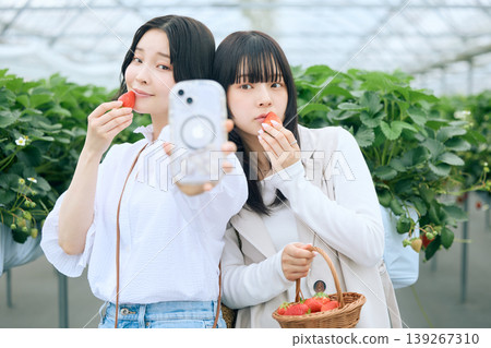 兩名年輕女子在草莓採摘溫室裡用智慧型手機自拍，她們手持草莓，笑容燦爛，展現彼此的友誼。 139267310