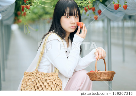 A young woman holds a freshly picked strawberry to her cheek at a strawberry picking greenhouse – a spring leisure experience. A young woman holds a freshly picked strawberry to her cheek at a strawberry picking greenhouse – a spring leisure experience. 139267315