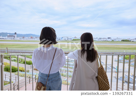 Two women looking out at the runway from an airport observation deck – an image of friends traveling or sightseeing. 139267331